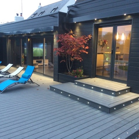 Ambiance de fin de journée sur une terrasse composite grise rétroéclairée par des spots LED intégrés, avec un salon de jardin moderne.