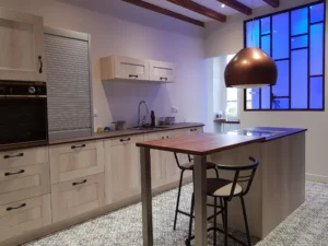 Cuisine en bois clair sur les façades de placard et les portes. Plan de travail en bois foncé qui contraste avec le reste de la cuisine. Îlot central plus petit mais très fonctionnel.