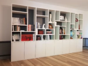 Vue d'ensemble d'un grand meuble bibliothèque blanc installé le long d'un mur, avec des niches décoratives colorées en gris et bleu pastel, surmontant des placards bas fermés.