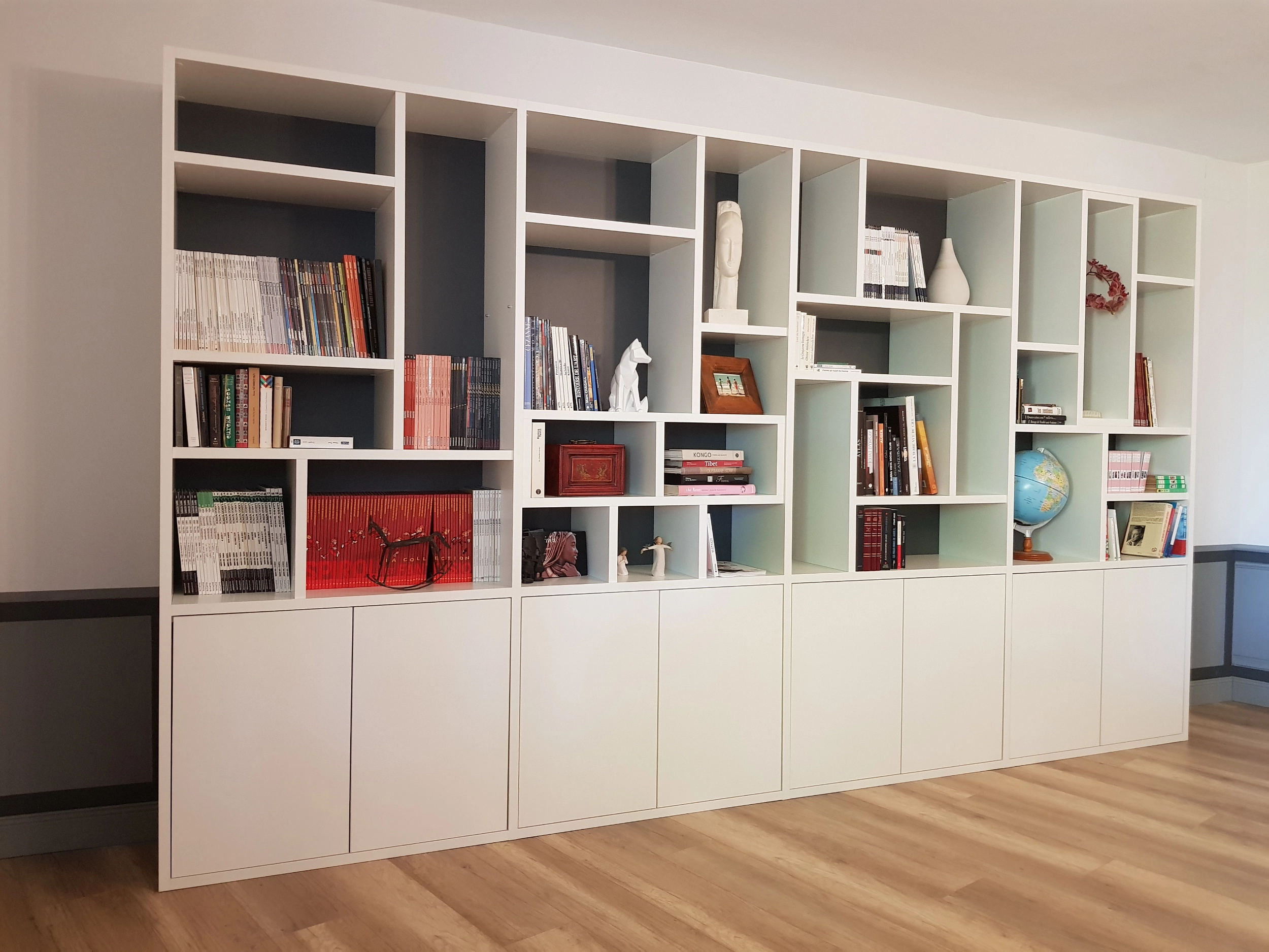 Vue d'ensemble d'un grand meuble bibliothèque blanc installé le long d'un mur, avec des niches décoratives colorées en gris et bleu pastel, surmontant des placards bas fermés.