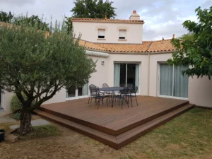Vue d'ensemble d'une grande terrasse surélevée en bois composite marron avec une table, des chaises et un olivier au premier plan devant une maison moderne.