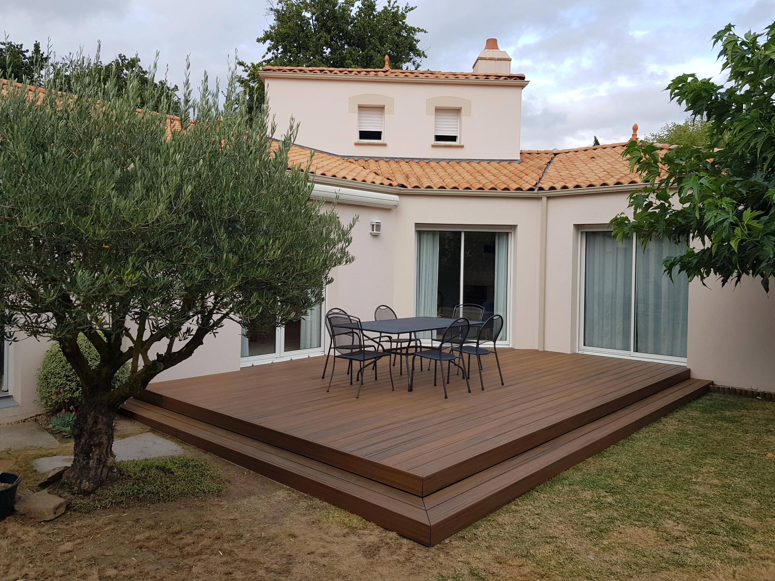 Vue d'ensemble d'une grande terrasse surélevée en bois composite marron avec une table, des chaises et un olivier au premier plan devant une maison moderne.