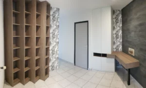 Vue d'angle montrant la bibliothèque en bois, la porte d'entrée grise, le placard blanc intégré et le bureau suspendu devant le mur d'ardoise noire.