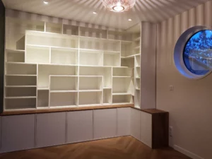 Grande bibliothèque blanche sur mesure épousant l'angle d'une pièce, dotée d'un long plateau de finition en bois chaud et surmontée de spots intégrés au plafond.