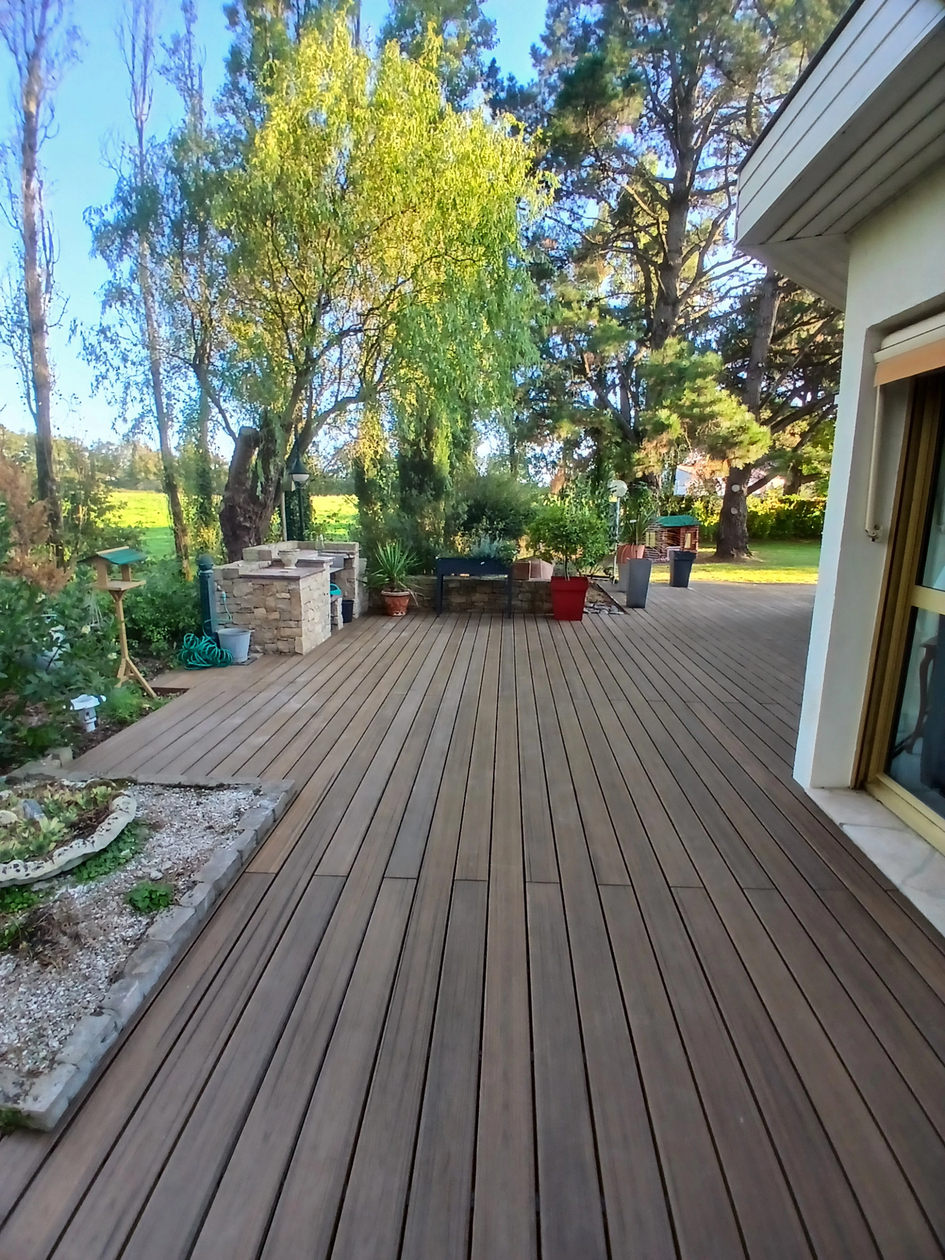 Longue terrasse en lames de composite marron longeant le jardin, avec un espace cuisine extérieure en pierre en arrière-plan.