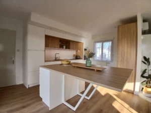Cuisine moderne blanche avec plan de travail en bois. Îlot central très grand. Mur entier de placard, et niche pour mettre l'évier et plan de travail.