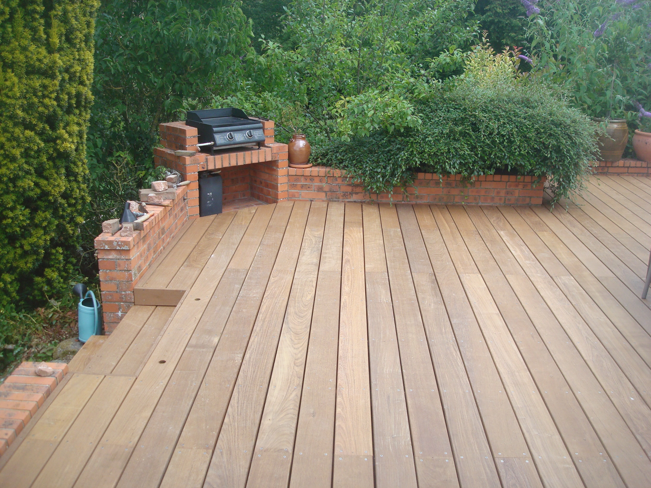 Vue de dessus d'une terrasse en lames de bois avec une structure de barbecue intégrée dans un muret en briques rouges.