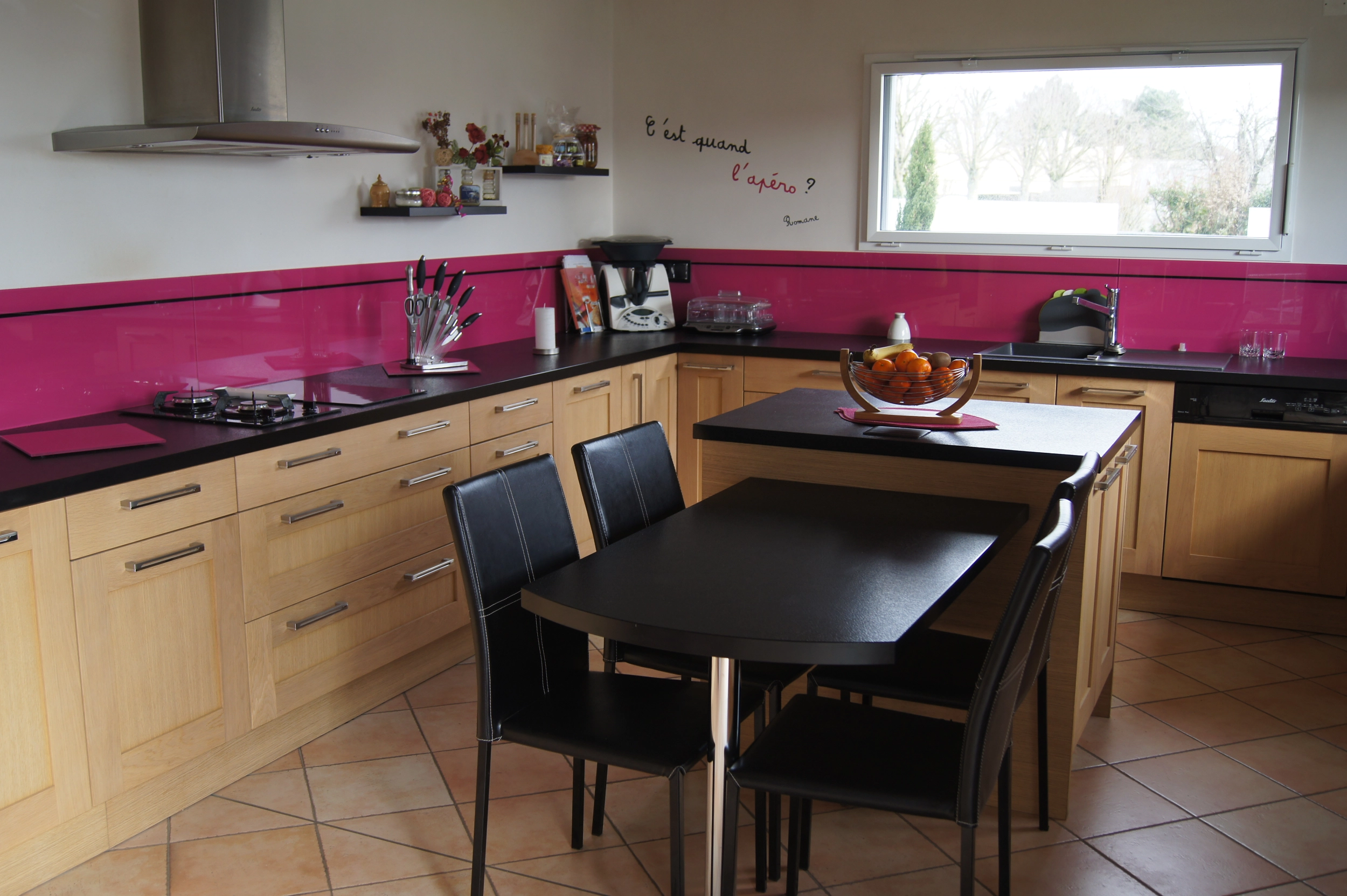 Cuisine en forme de L avec beaucoup de placard. Un grand plan de travail tout le long du L de la cuisine. Crédence de couleur rose.