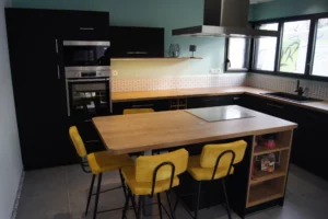 Cuisine en L avec un grand plan de travail en bois. Les façades de placard et les portes sont noires mates. Un îlot central avec des places pour le déjeuner, la plaque de cuisson et des rangements. Grande cuisine familiale.