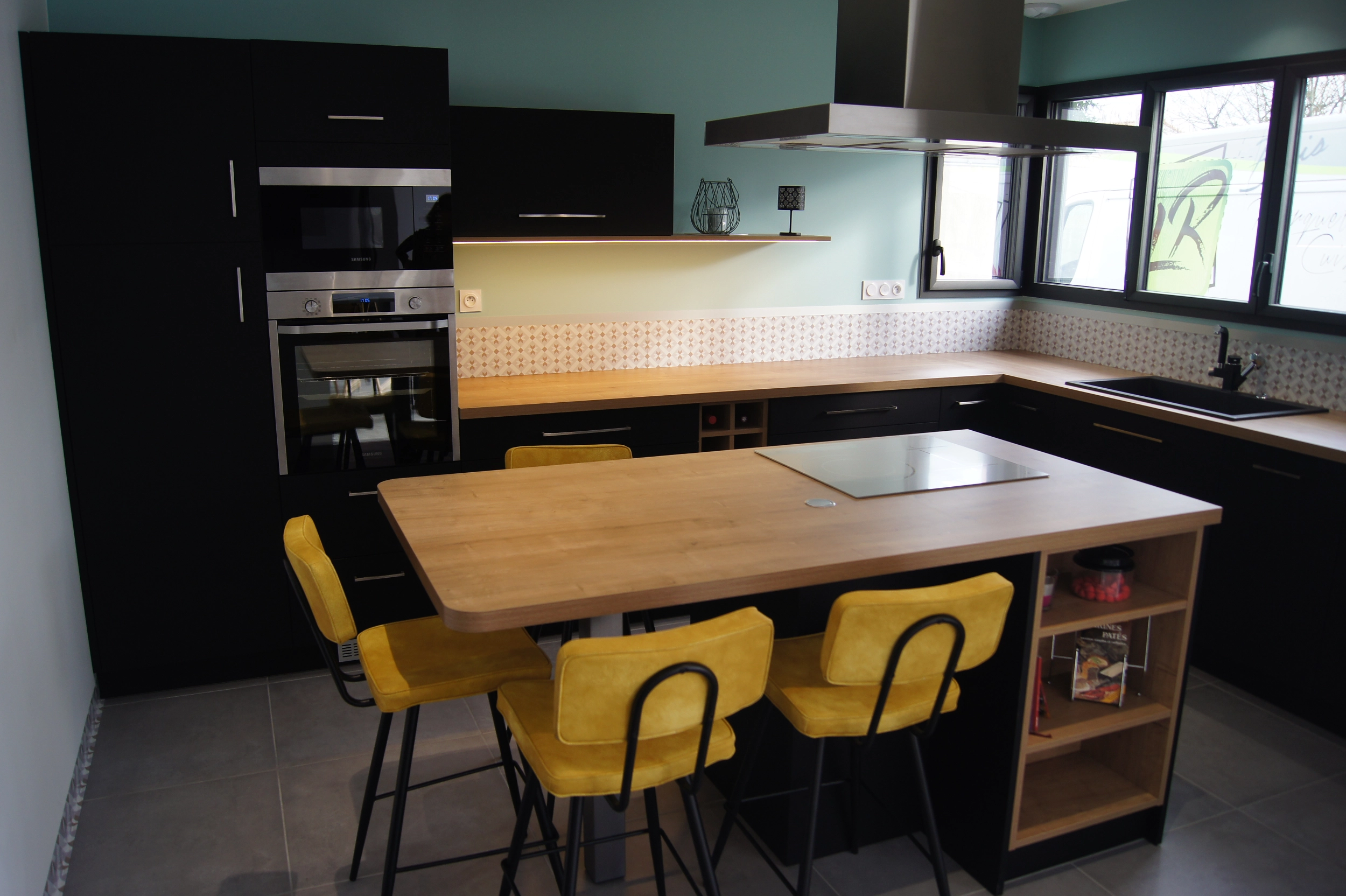 Cuisine en L avec un grand plan de travail en bois. Les façades de placard et les portes sont noires mates. Un îlot central avec des places pour le déjeuner, la plaque de cuisson et des rangements. Grande cuisine familiale.