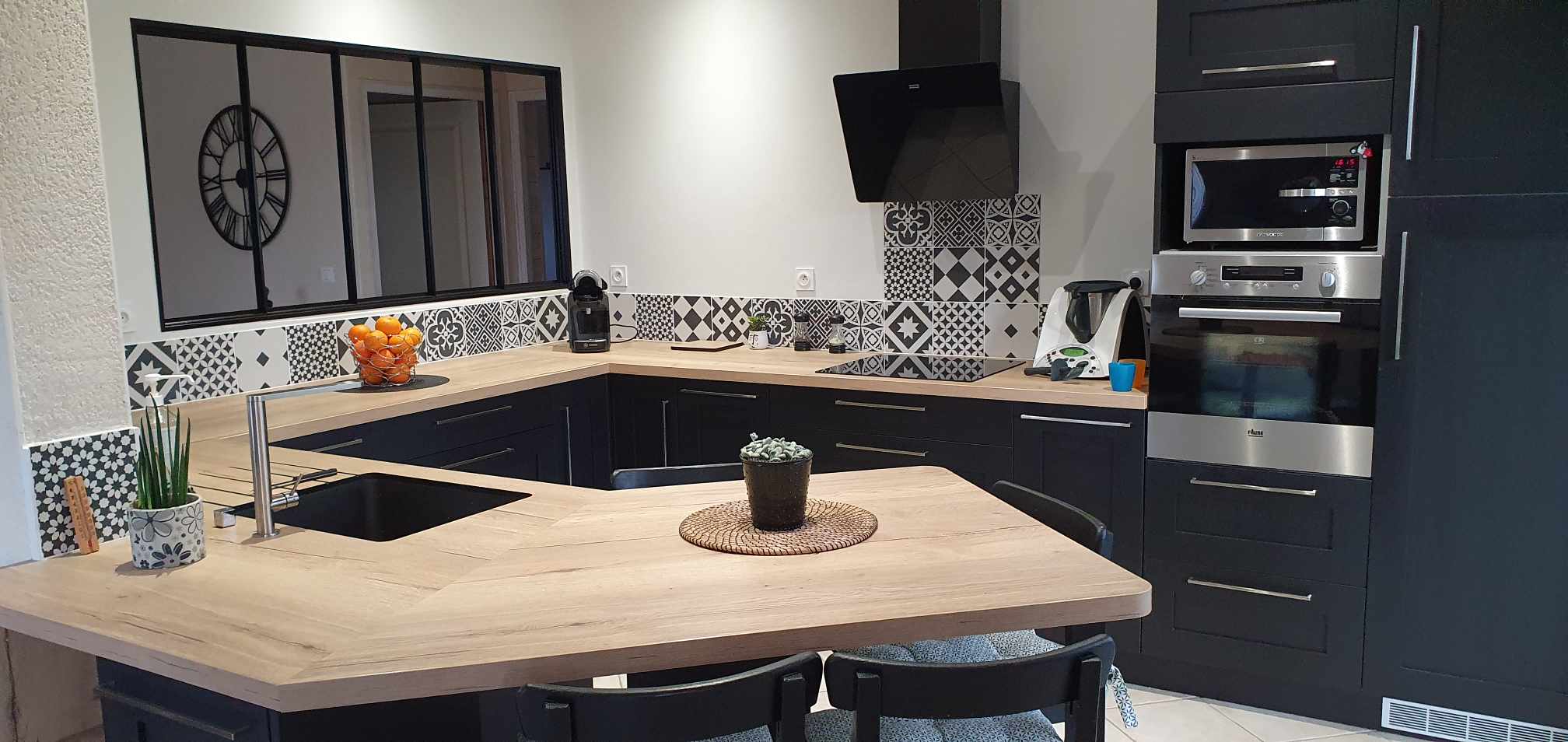 Cuisine moderne en U avec meubles noirs mats, plan de travail en chêne et crédence en carreaux de ciment.